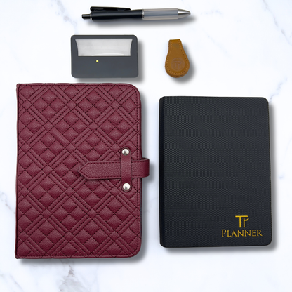 A styled flat-lay of stationery accessories on a white marble background, featuring a burgundy quilted planner cover with a strap closure, a black textured planner, a black pen, a small black card holder, and a brown leather keychain.