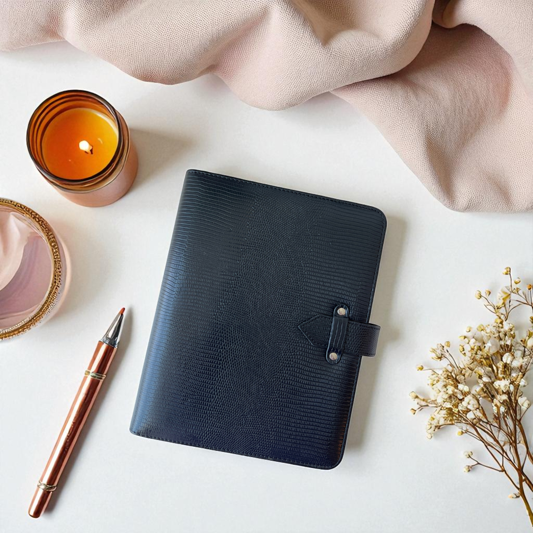Black planner on a white surface with a candle, pen, and flowers.