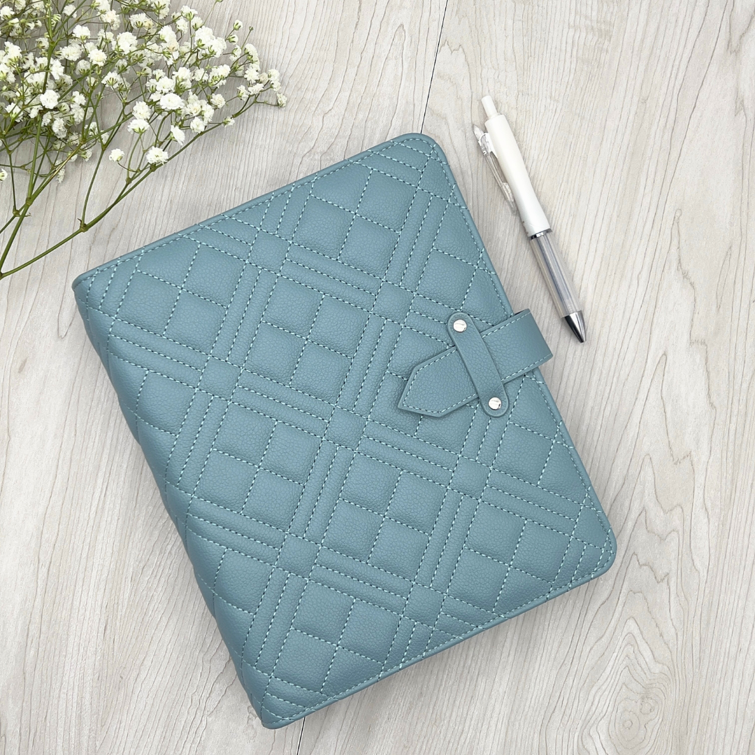 Blue quilted planner on a light wooden surface with a pen and flowers.