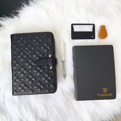 Black planner with textured cover, pen, card magnifier, and magnetic clip on a white fur surface